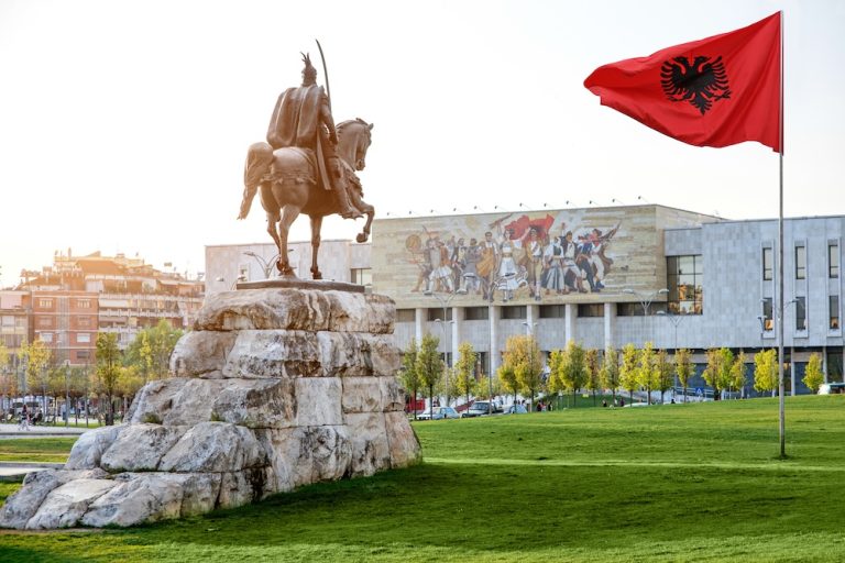 Estátua de Skanderbeg e bandeira da Albânia em Tirana, com edifício histórico ao fundo – viagem à Albânia e Montenegro